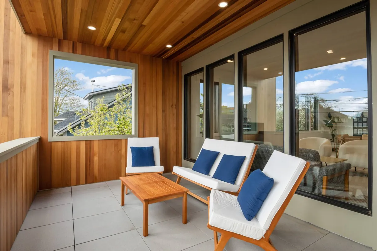 A tranquil covered patio featuring a wooden ceiling, comfortable seating arrangements, and large windows that invite plenty of natural light. The space is styled for relaxation, making it perfect for lounging, reading, or enjoying a cup of coffee while taking in the ambiance of the neighborhood.