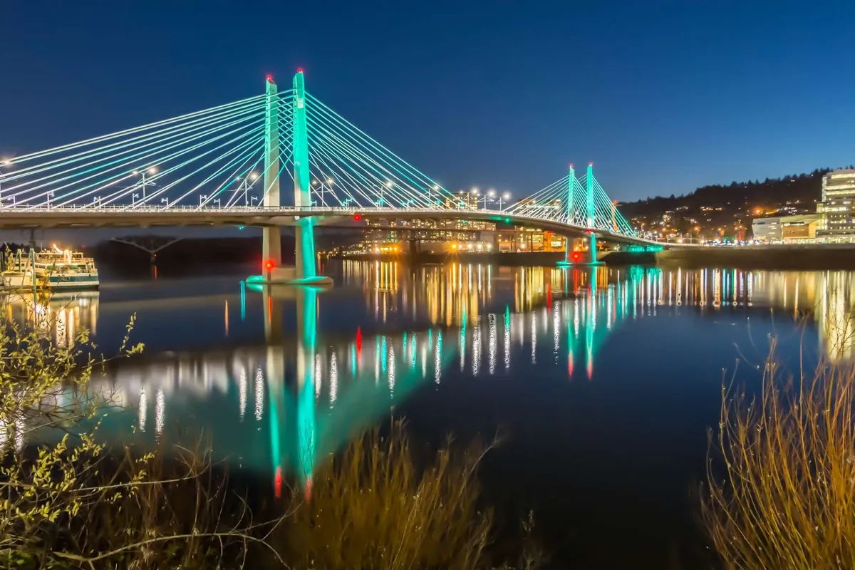 A serene evening shot of a suspension bridge spanning the river, with city lights reflecting in the water. The scene is lively yet peaceful, evoking the spirit of riverside walks and the city’s vibrant nightlife.