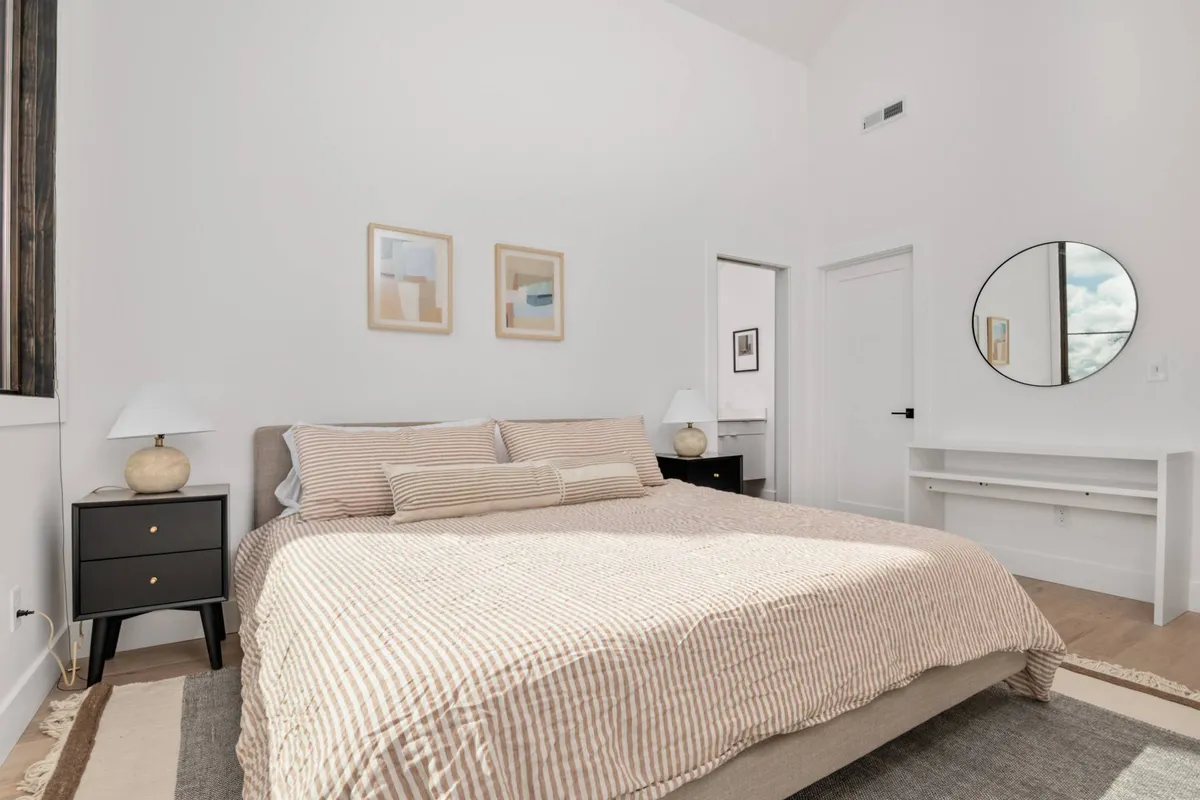 A tranquil bedroom scene featuring a plush bed framed by nightstands and calming artwork. A large mirror reflects the soft, natural light, enhancing the sense of space and comfort. The setting exudes an inviting, peaceful mood, ideal for relaxation and restorative sleep.