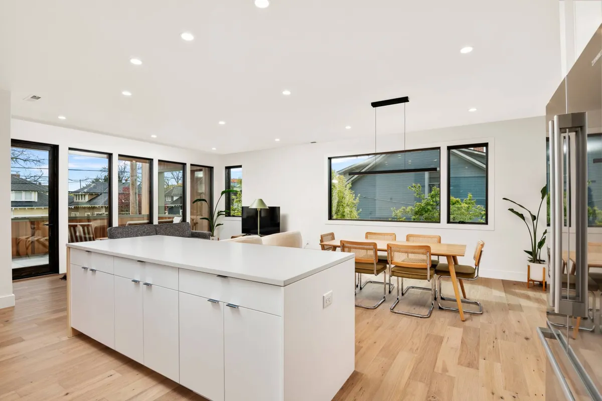 This image showcases an open-plan layout with an island counter, dining table, large windows, and a seating area. The abundance of natural light and airy arrangement embodies the social, Scandinavian-inspired atmosphere emphasized in the section. Several communal spaces, including living, dining, and kitchen, are visible in a single frame.