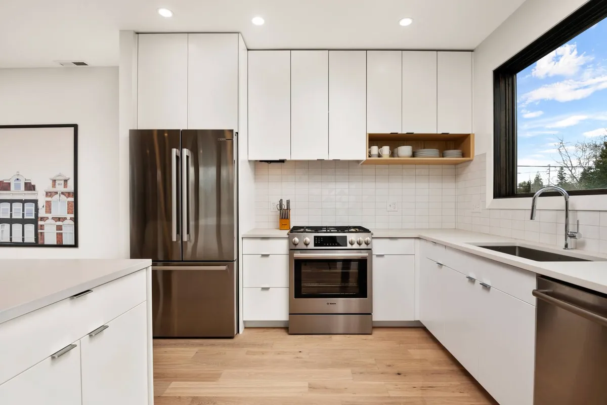 A modern kitchen scene with a stainless steel refrigerator, contemporary stove, open shelving with dishes on display, and a large window letting in ample daylight. The kitchen is designed for cooking, hosting, and gathering.