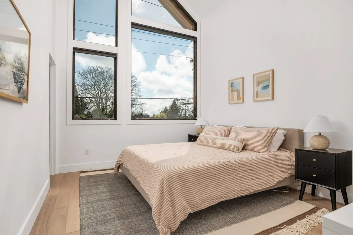 A serene bedroom filled with natural light from expansive windows. The decor is minimalist yet cozy, with a plush bed, nightstands, and understated framed artwork adding to the tranquil, Scandinavian-inspired aesthetic. Soft bedding and calming hues create an inviting, restful atmosphere ideal for a private retreat.
