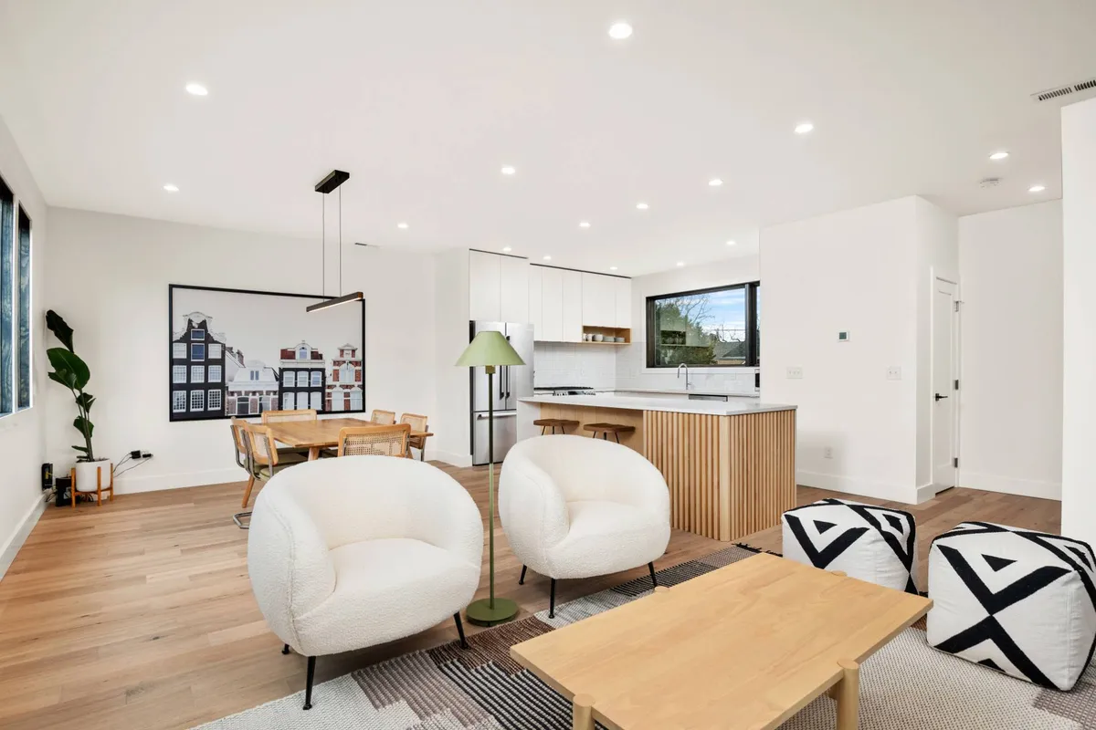 A stylish interior view showing a welcoming living area with sofa chairs, a dining table, and the kitchen area in the background. Artwork on the wall and natural light reinforce a contemporary, inviting atmosphere suited for both relaxation and productivity.