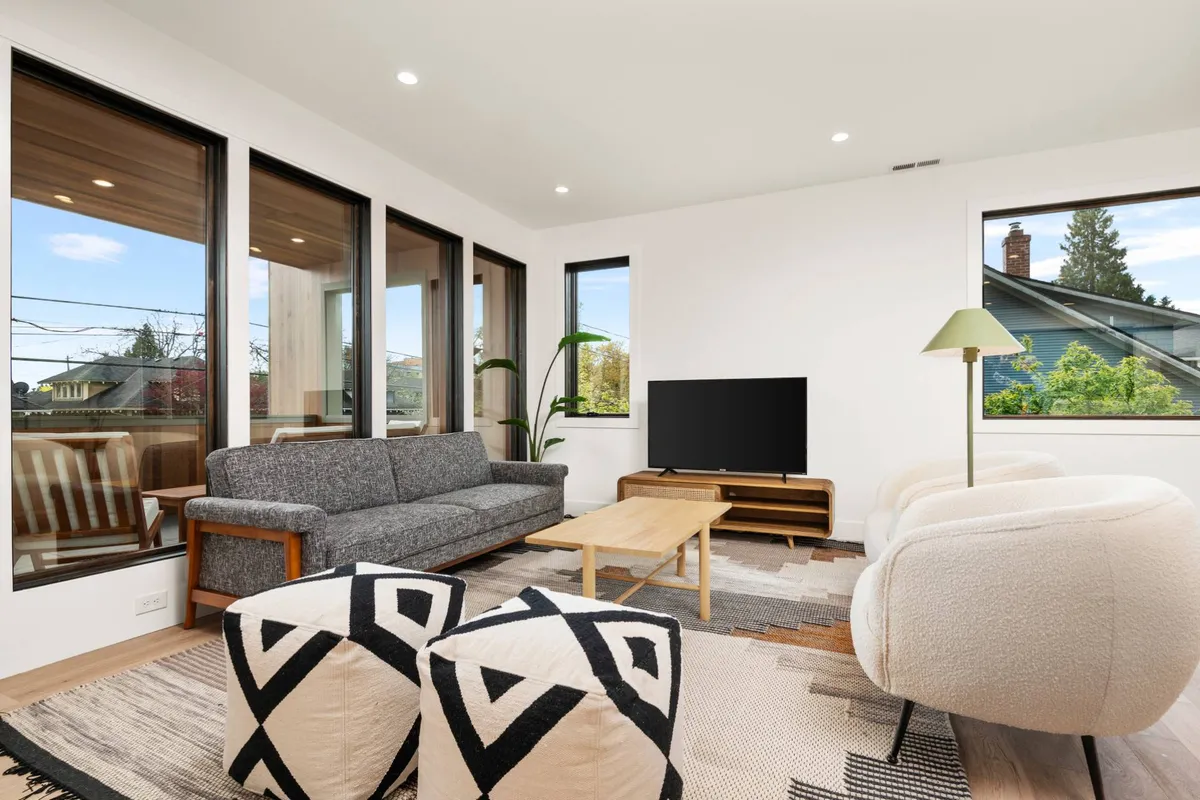 A bright, welcoming living area featuring a sofa, coffee table, armchairs, television, and expansive windows. The room is bathed in sunlight and set up for relaxing, reading, or entertaining.